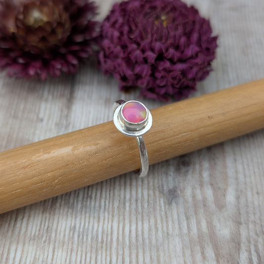 Handmade sterling silver stacking ring with a pink aurora opal cabochon, displayed on a wooden ring mandrel with dried purple flowers in the background.