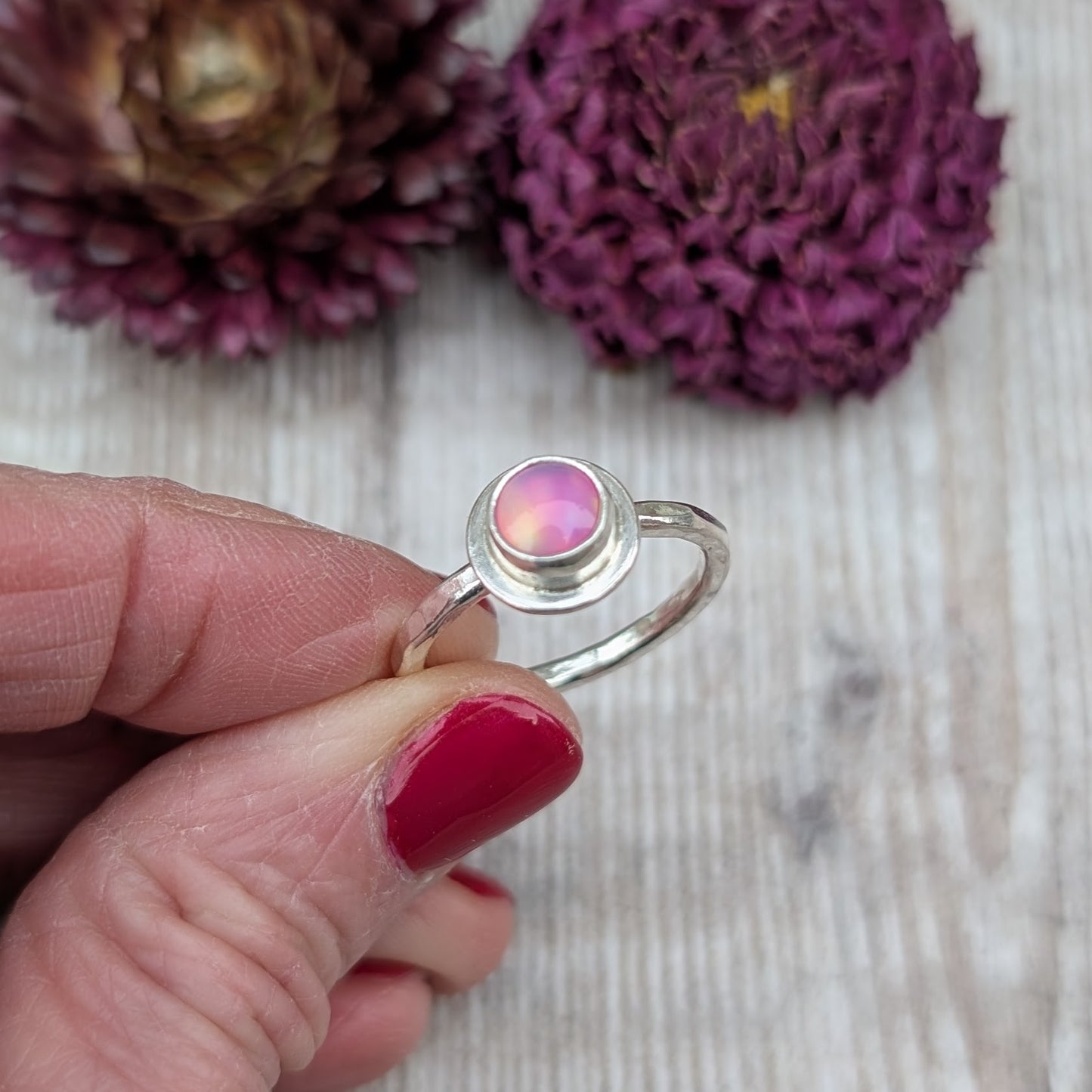 Close-up of a handmade sterling silver ring held between fingers, featuring a round pink aurora opal set in a smooth silver bezel.