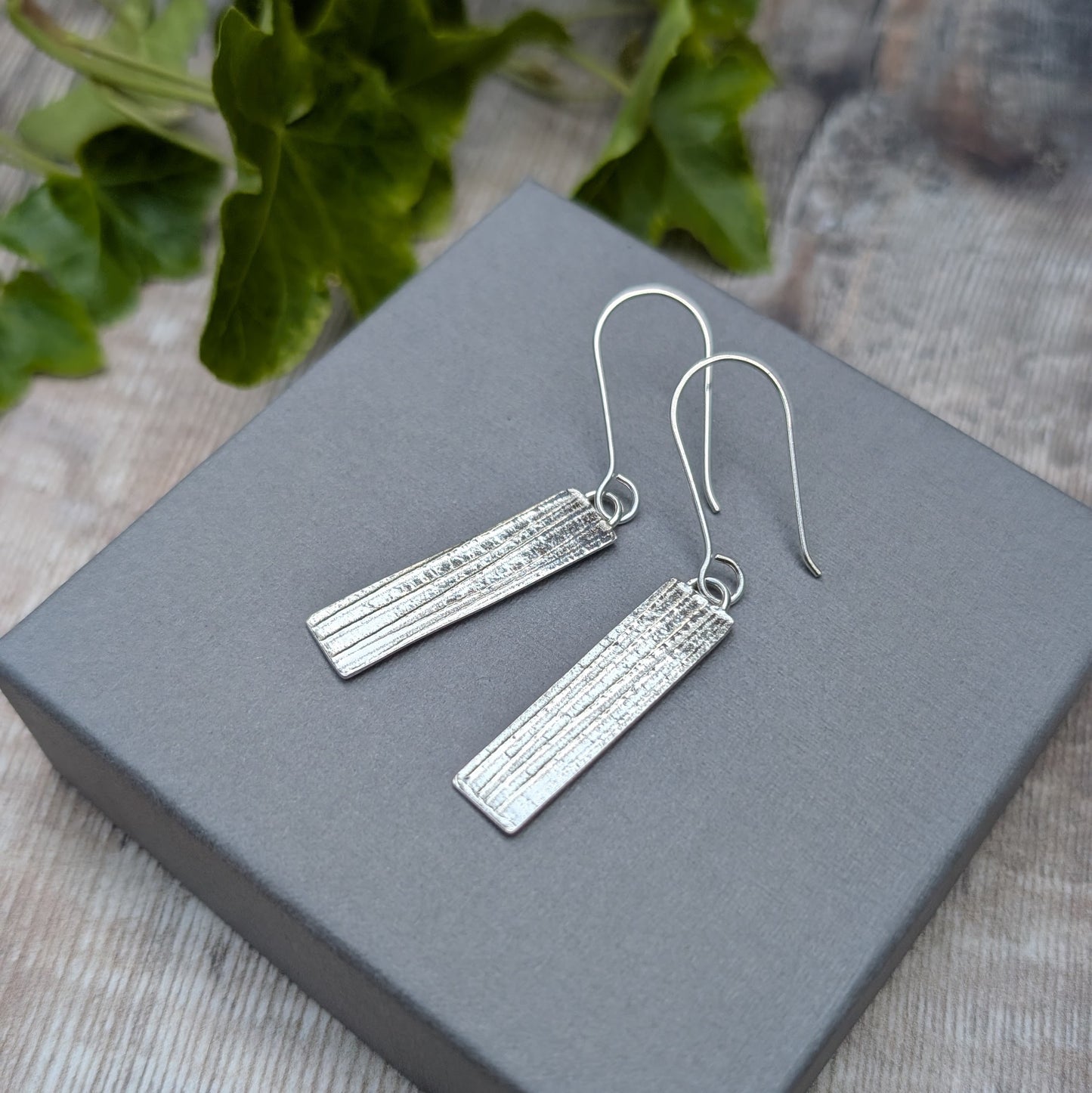 Silver Rectangle Earrings with Ribbon Pattern on a grey box with green leaves in the background
