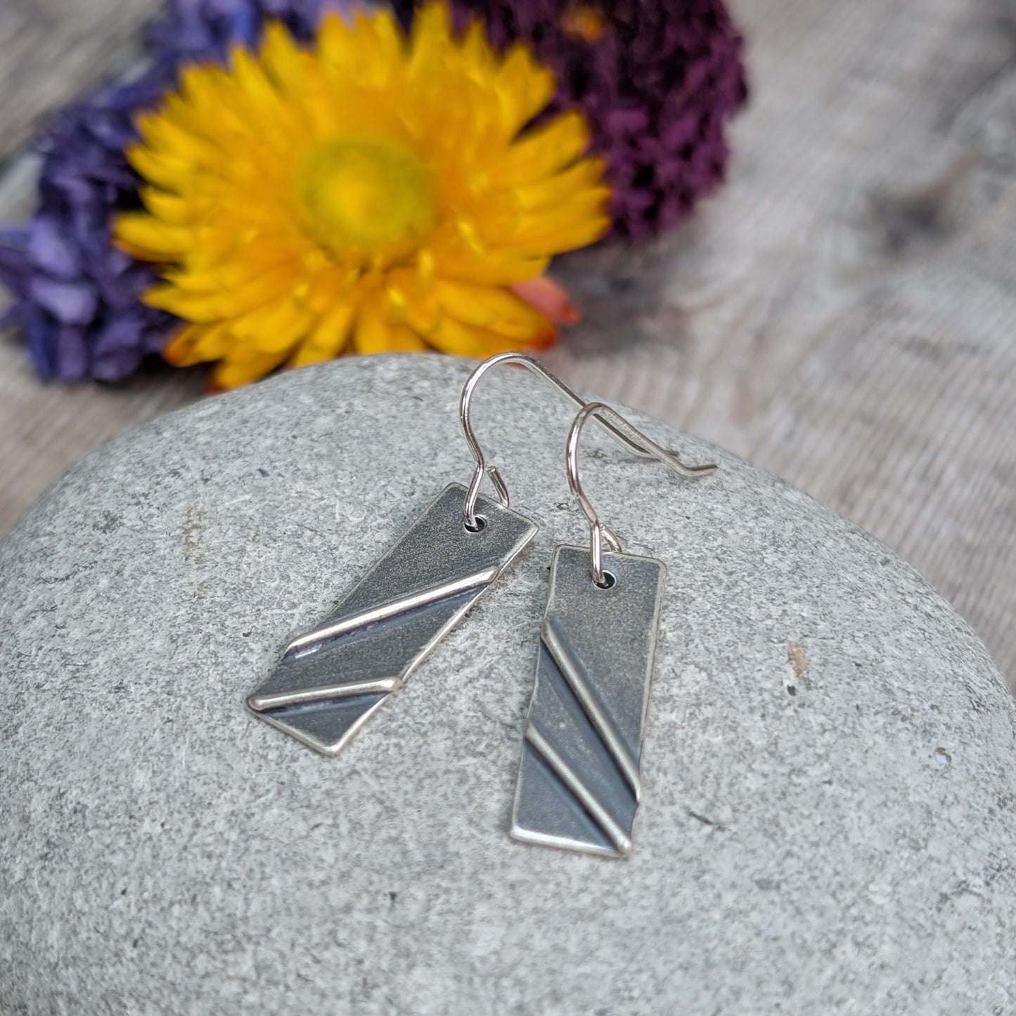 Each earring is a silver rectangle shape. Two raised metal lines are set on top, spaced apart and set at diagonals. Silver finish is slightly textured and oxidised to enhance the raised details. Suspended from silver earring wire.