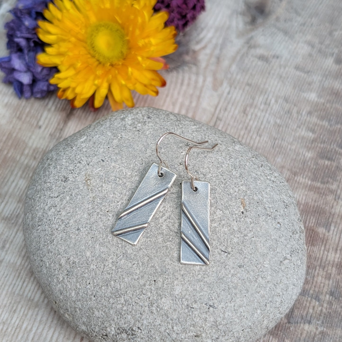 Each earring is a silver rectangle shape. Two raised metal lines are set on top, spaced apart and set at diagonals. Silver finish is slightly textured and oxidised to enhance the raised details. Suspended from silver earring wire.