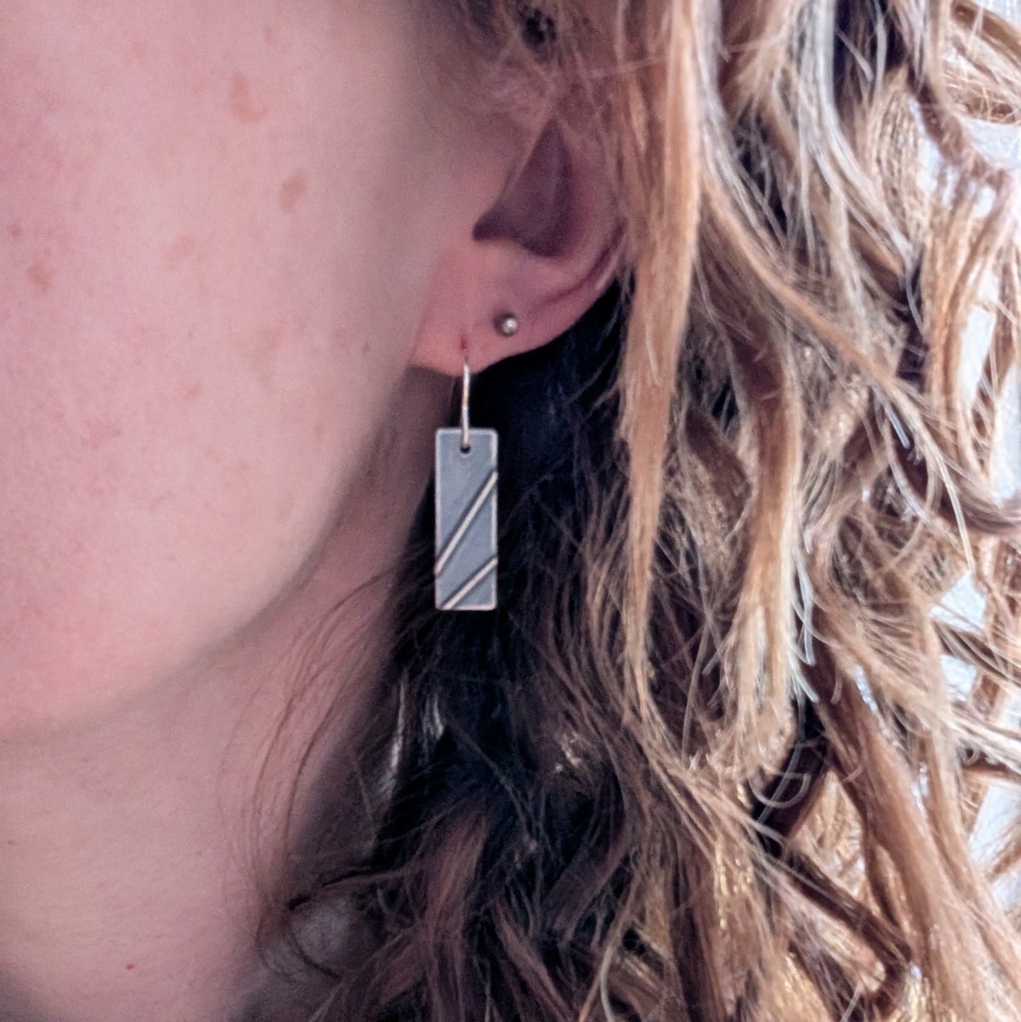 Each earring is a silver rectangle shape. Two raised metal lines are set on top, spaced apart and set at diagonals. Silver finish is slightly textured and oxidised to enhance the raised details. Suspended from silver earring wire.