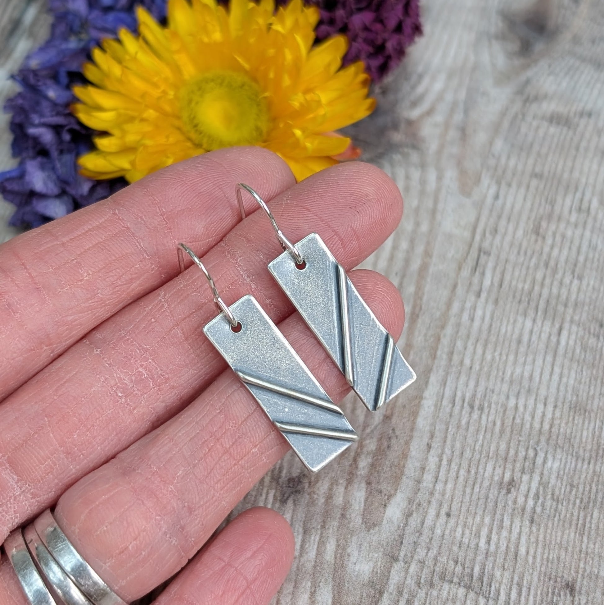 Each earring is a silver rectangle shape. Two raised metal lines are set on top, spaced apart and set at diagonals. Silver finish is slightly textured and oxidised to enhance the raised details. Suspended from silver earring wire.