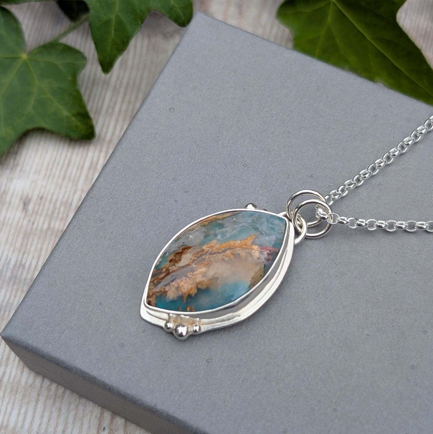 Silver Regency Rose Plume Agate Necklace photographed on a grey jewellery box with ivy leaves in the background, highlighting the natural colours and handmade Sterling Silver setting.
