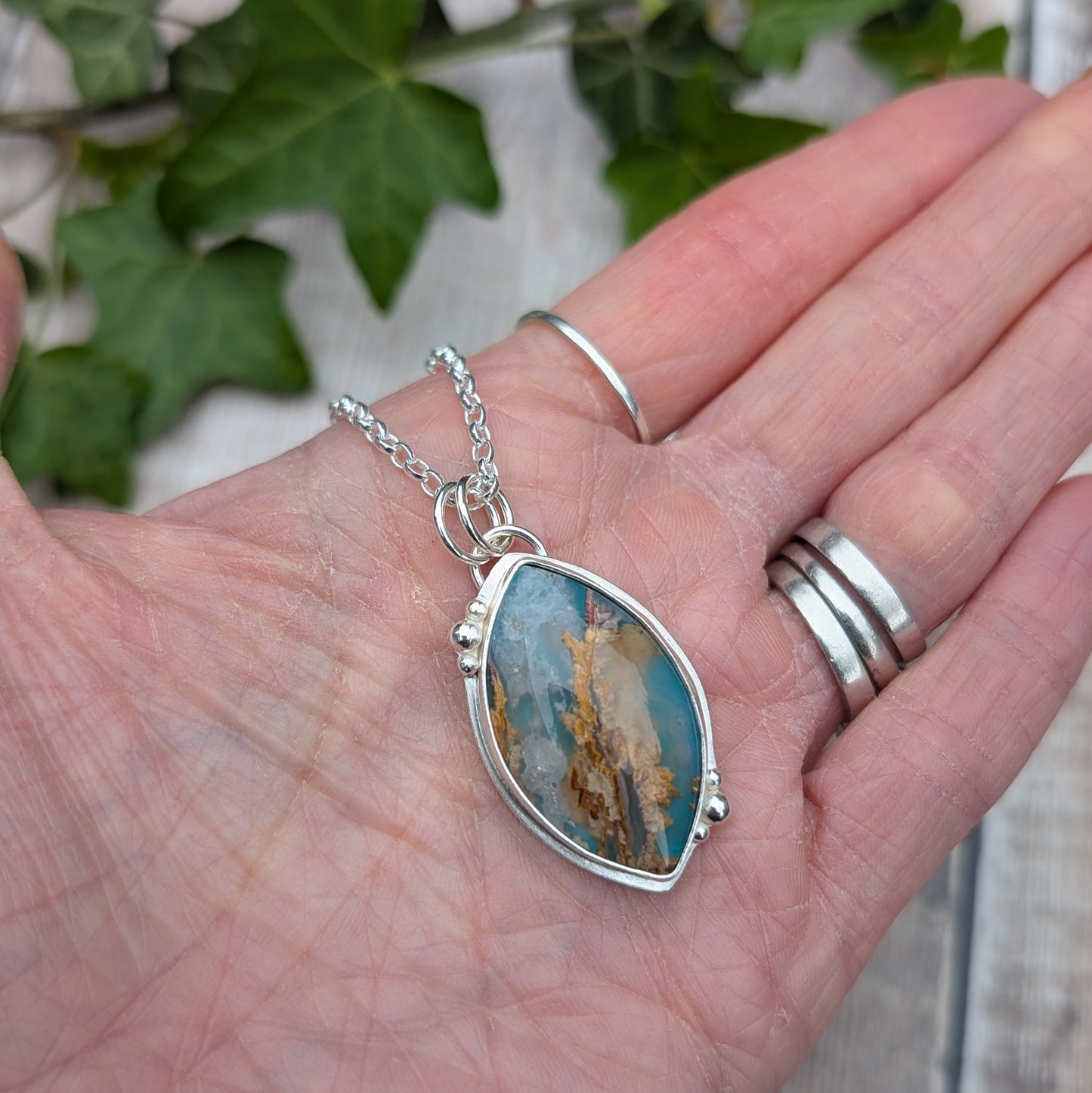 Silver Regency Rose Plume Agate Necklace resting in a hand, showing the handmade recycled Sterling Silver setting and natural blue and earthy patterned stone.