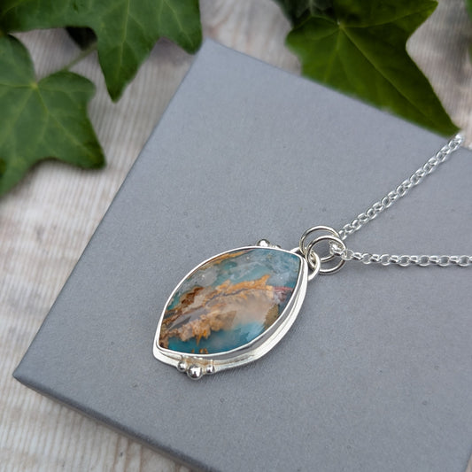 Close-up of a Silver Regency Rose Plume Agate Necklace on a grey jewellery box, showing the natural blue, cream and earthy patterns in the stone and handmade silver setting.