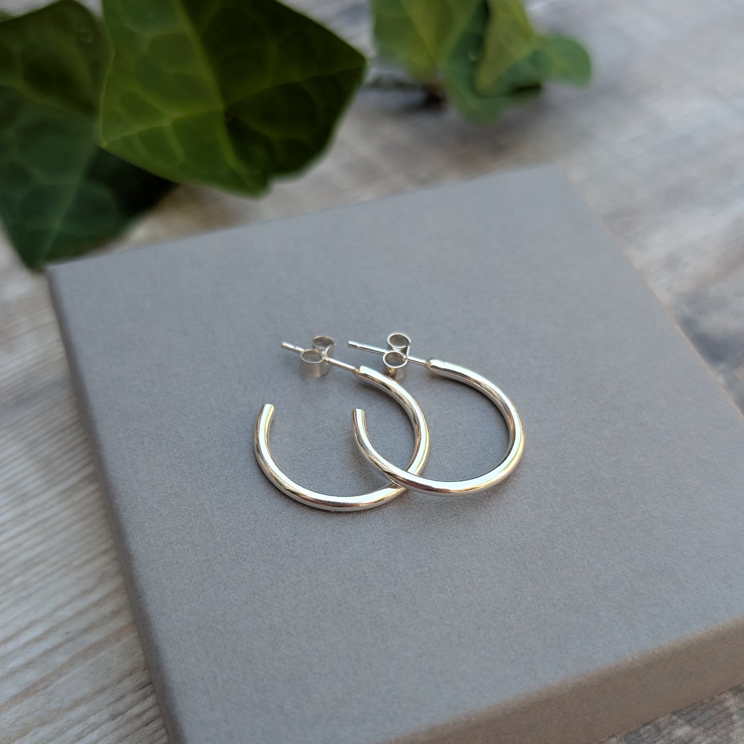Silver smooth hoop earrings handmade from recycled Sterling Silver shown on a grey jewellery box with green leaves in the background.