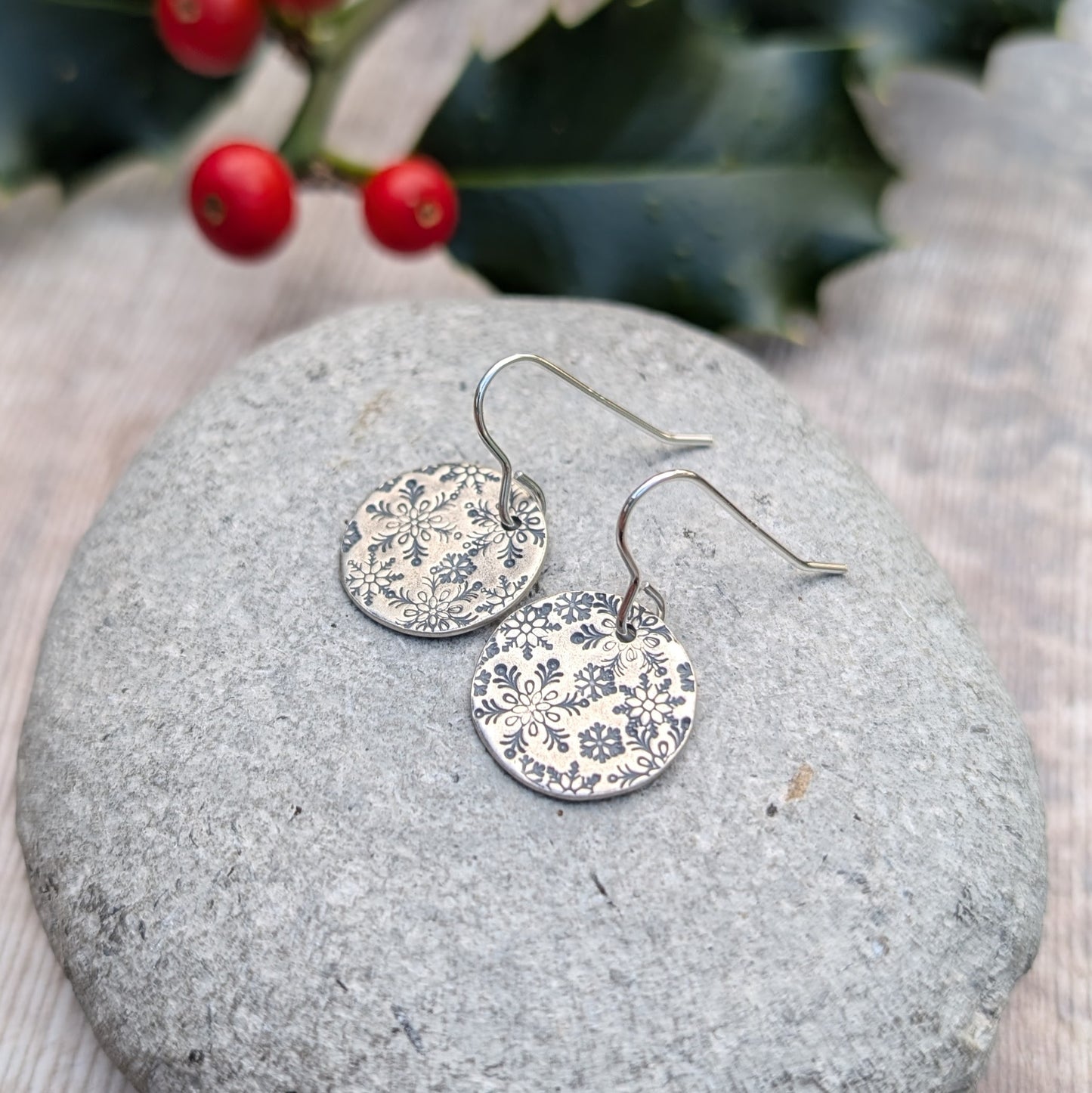 A pair of oxidised silver snowflake disc earrings resting on a smooth grey stone, highlighting the intricate winter-inspired snowflake patterns, with festive holly and berries in the background.