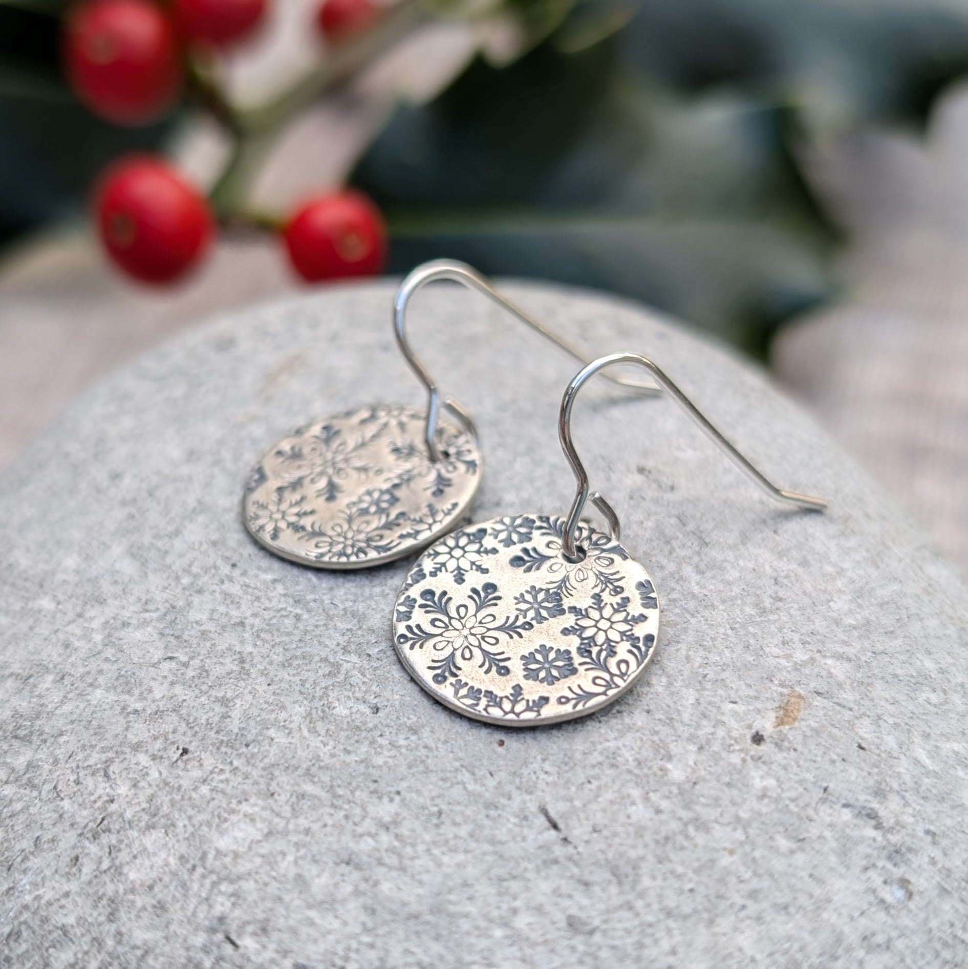 A pair of oxidised silver snowflake disc earrings resting on a smooth grey stone, highlighting the intricate winter-inspired snowflake patterns, with festive holly and berries in the background.