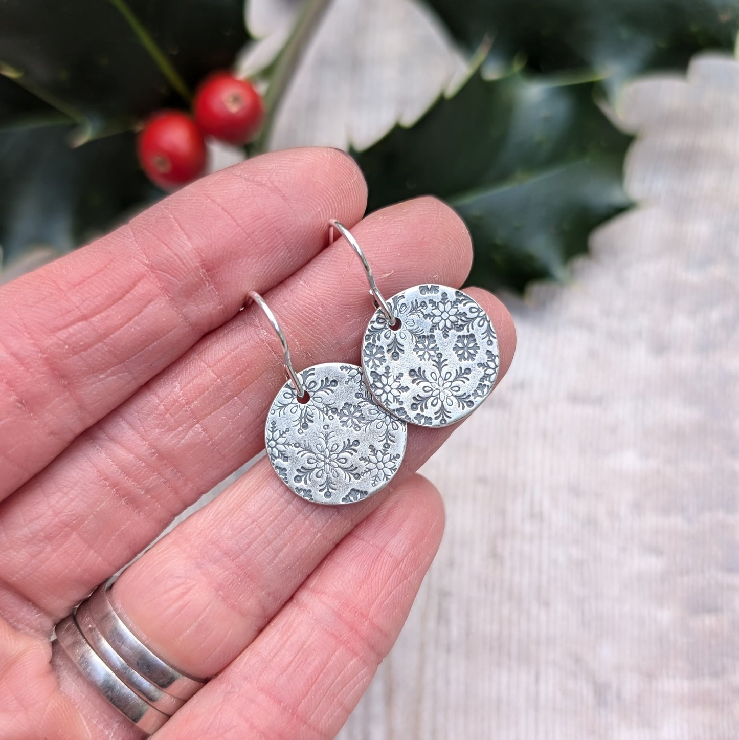 Close-up of handmade oxidised silver snowflake disc earrings, featuring intricate snowflake patterns, held in hand with a festive holly branch and berries in the background.