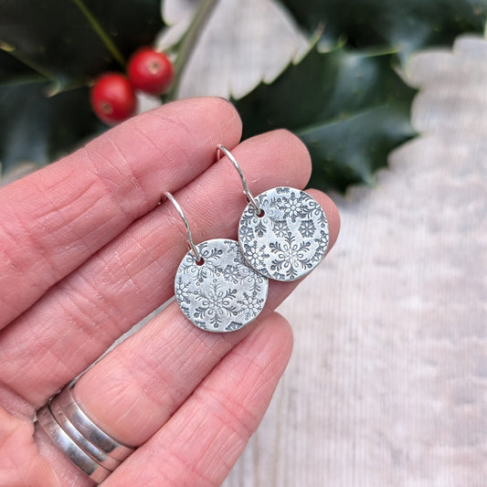 Close-up of handmade oxidised silver snowflake disc earrings, featuring intricate snowflake patterns, held in hand with a festive holly branch and berries in the background.