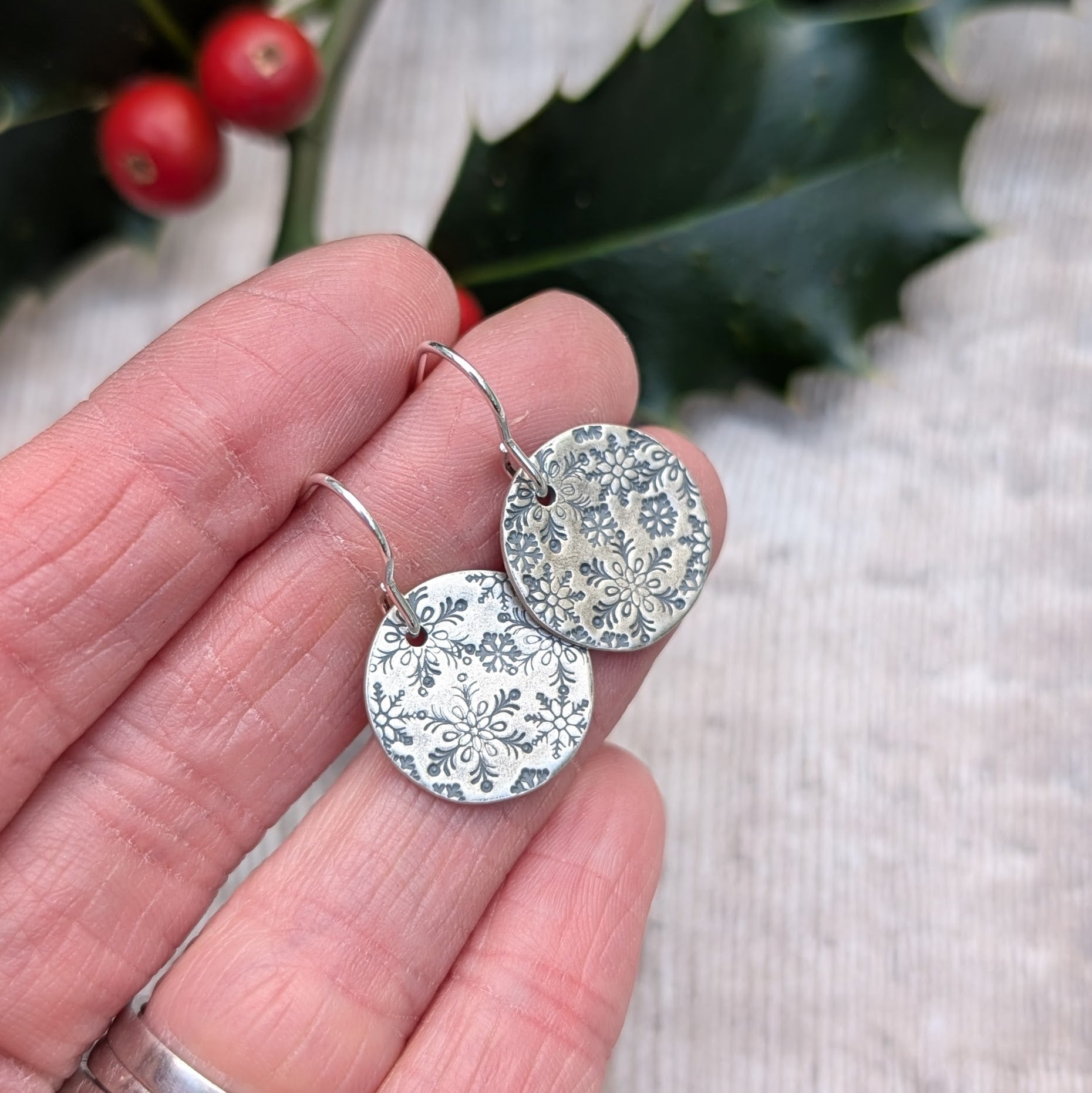 Close-up of handmade oxidised silver snowflake disc earrings, featuring intricate snowflake patterns, held in hand with a festive holly branch and berries in the background.