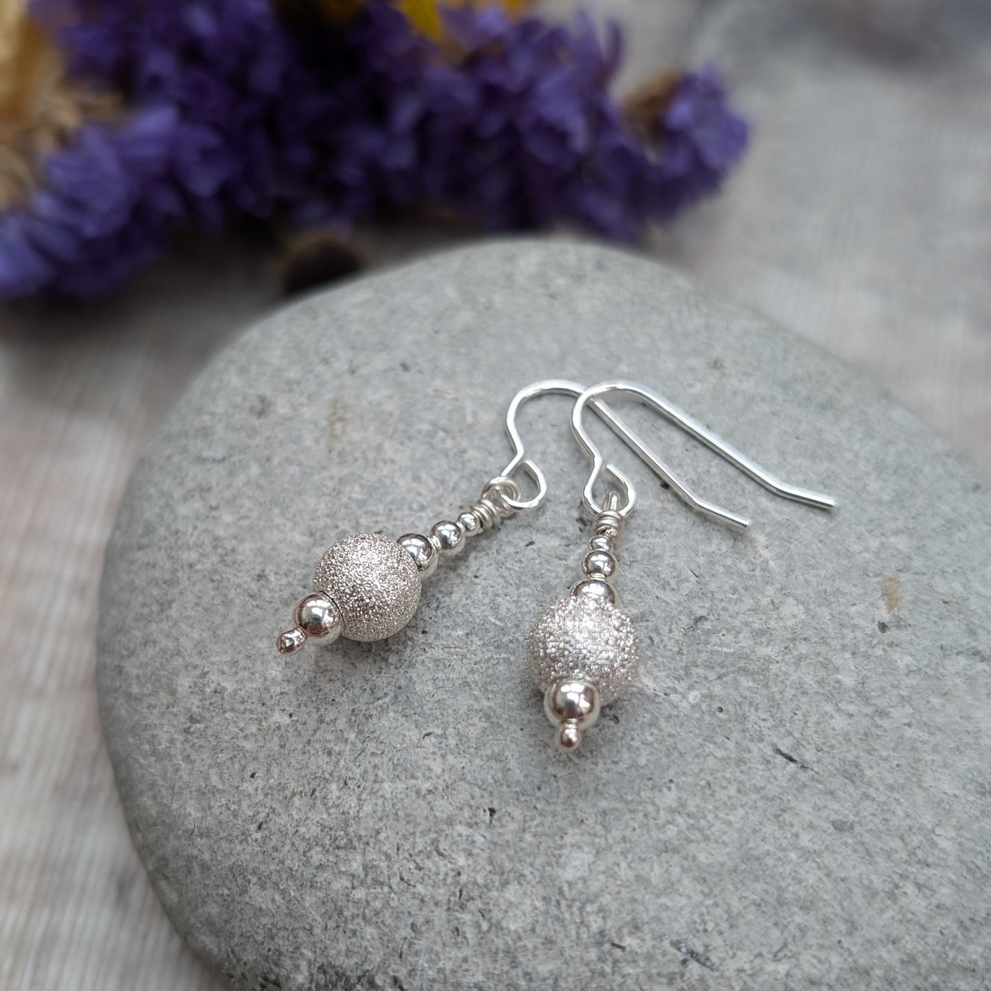 Pair of silver sparkle bead earrings resting on a smooth grey stone, with small polished beads and a central glittery bead catching the light, with blurred flowers in the background.