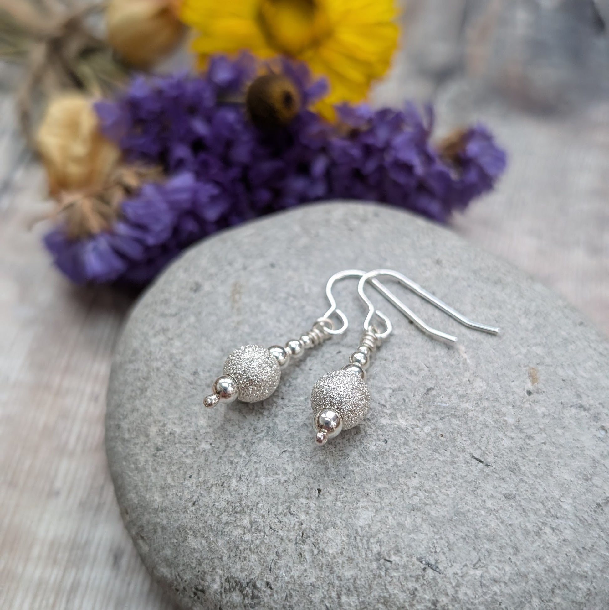 Pair of silver sparkle bead earrings resting on a smooth grey stone, with small polished beads and a central glittery bead catching the light, with blurred flowers in the background.