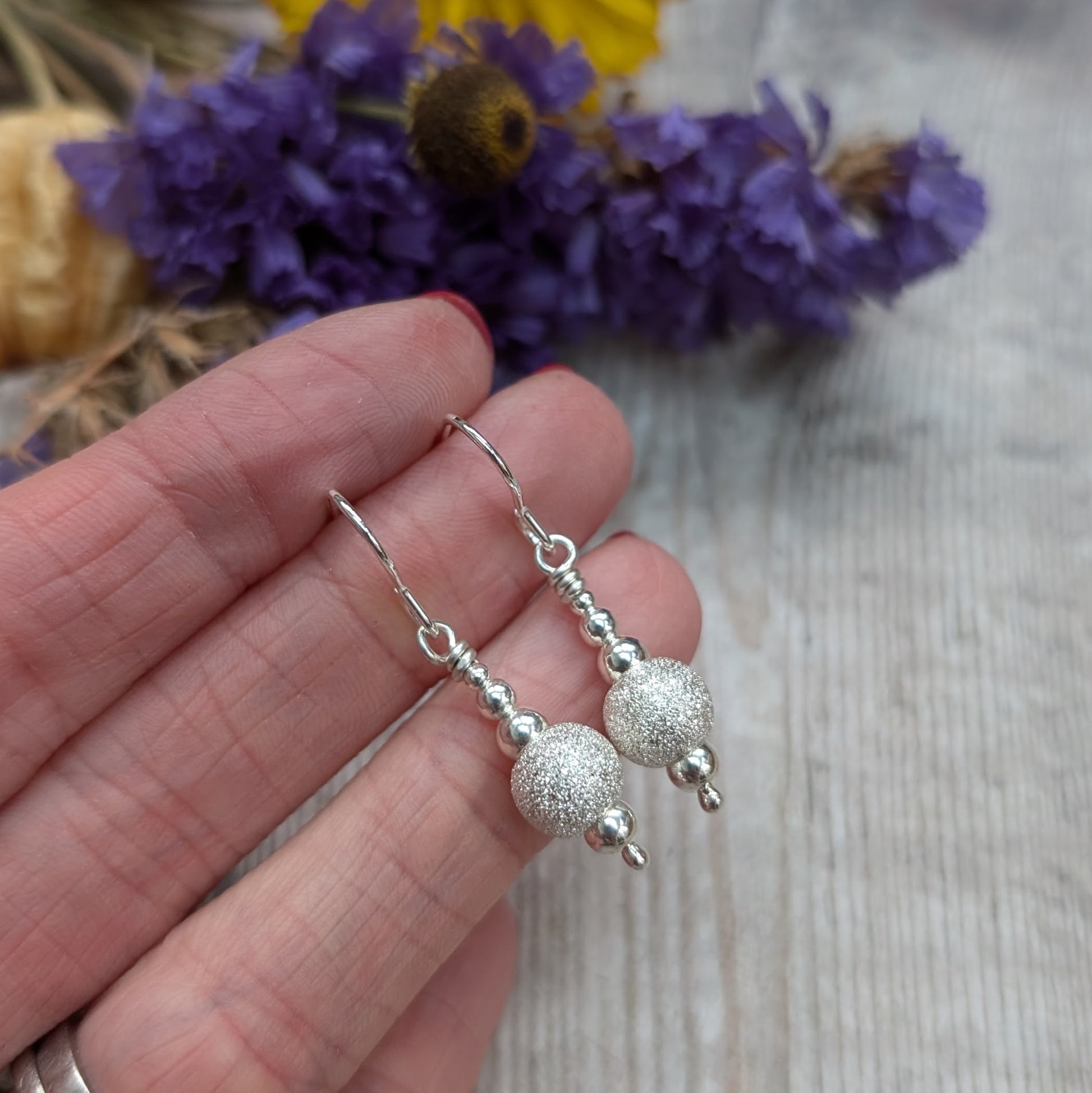 Hand holding a pair of silver sparkle bead earrings with hook fittings, featuring a glittery central bead and small polished silver beads above and below, with purple and yellow flowers blurred in the background.