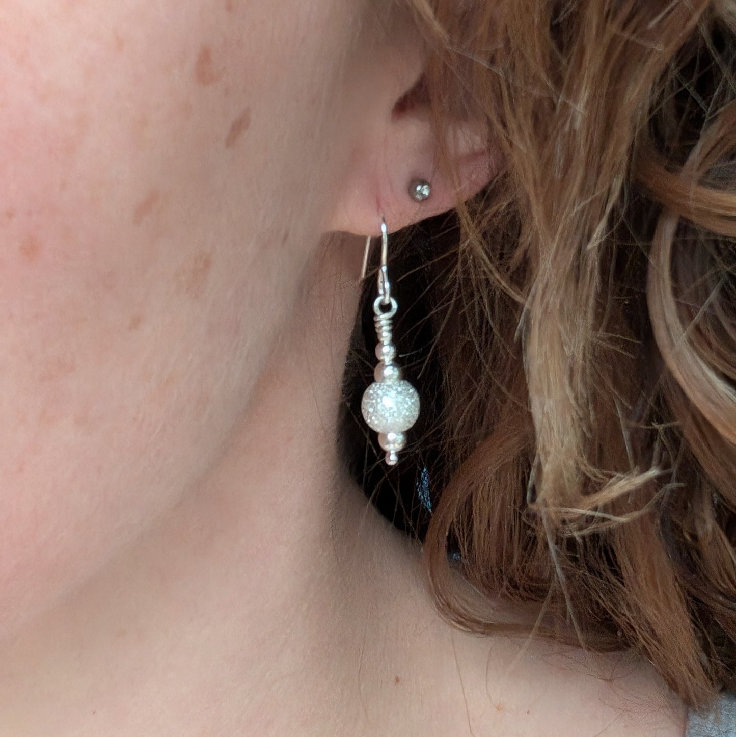Close-up of a person wearing one silver sparkle bead earring, showing the glittery bead detail hanging just below the earlobe.