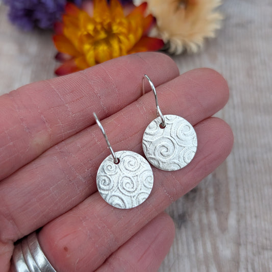 Silver Spiral Pattern Disc Earrings close up in hand with flowers in the background, showing texture and finish