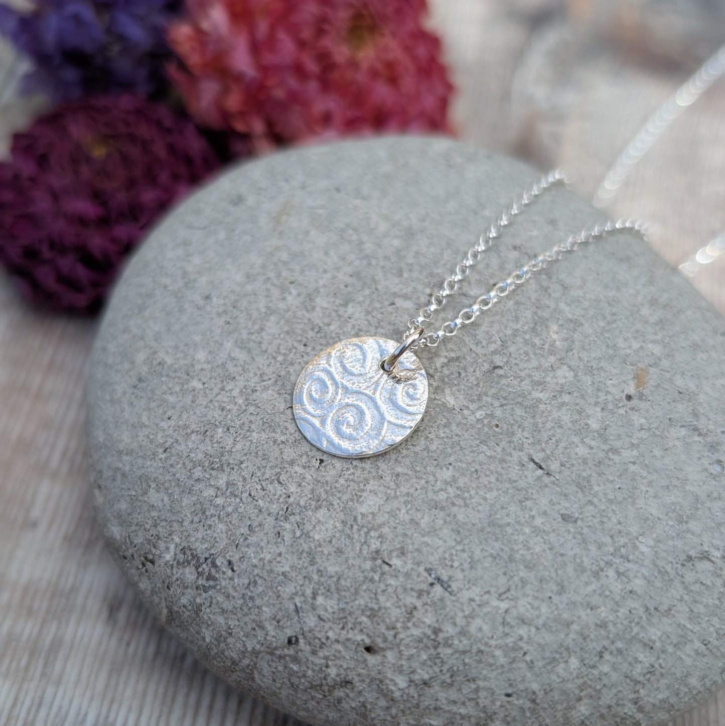 Silver Spiral Pattern Disc Necklace resting on a grey stone with dried pink and purple flowers in the background