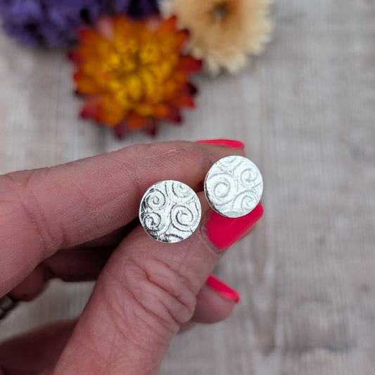 Silver Spiral Pattern Disc Stud Earrings close up in hand with flowers in the background, showing texture and finish