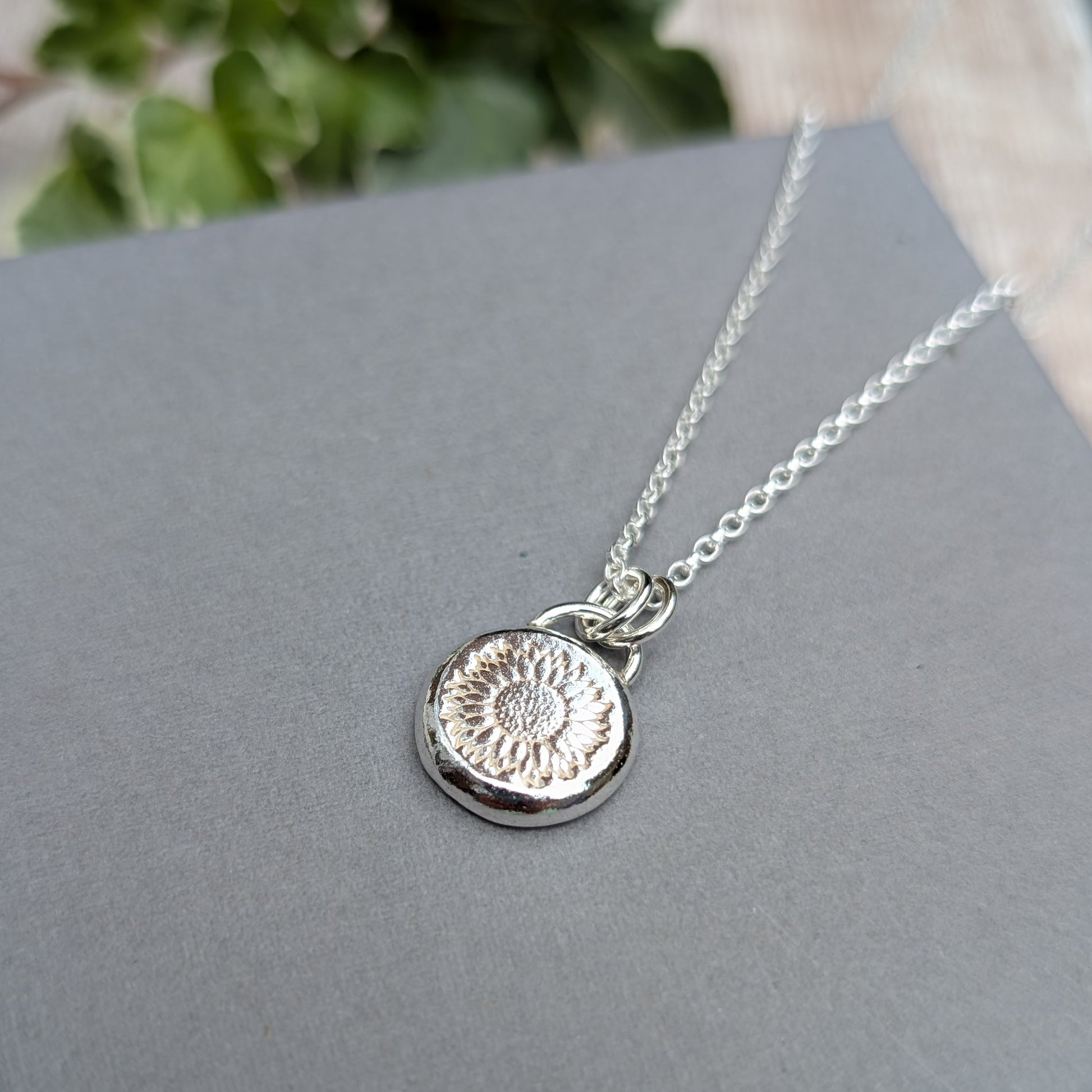 Close up of the handmade Silver Sunflower Pebble Necklace with stamped sunflower detail, displayed on a grey background with greenery behind.