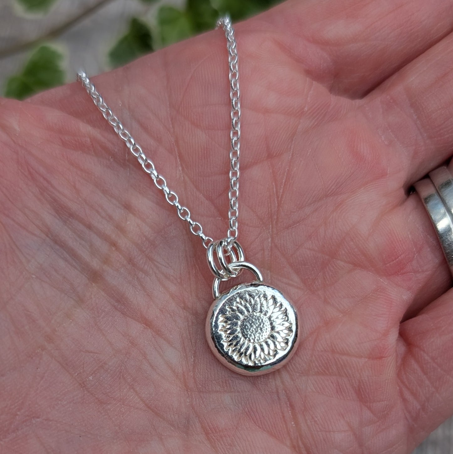 Close up of the Silver Sunflower Pebble Necklace held in a hand, showing the detailed sunflower stamped into the round silver pebble pendant.