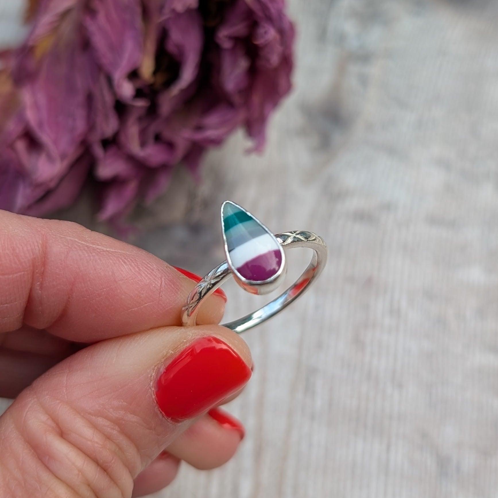 Close-up of a hand holding a silver ring with a striped teardrop-shaped Surfite stone and patterned band, in front of dried purple petals.