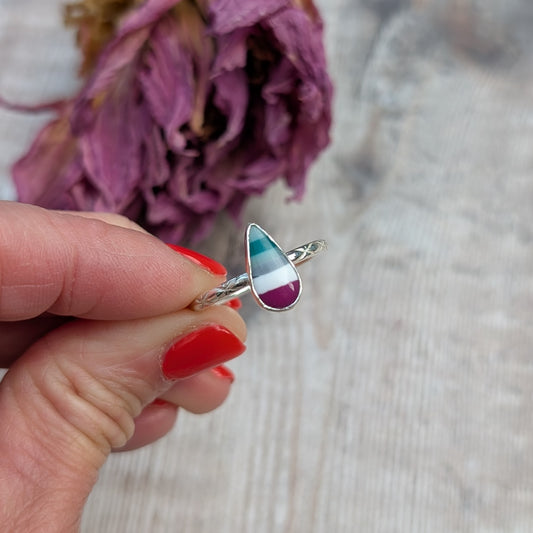 Close-up of a hand holding a silver ring with a striped teardrop-shaped Surfite stone and patterned band, in front of dried purple petals.