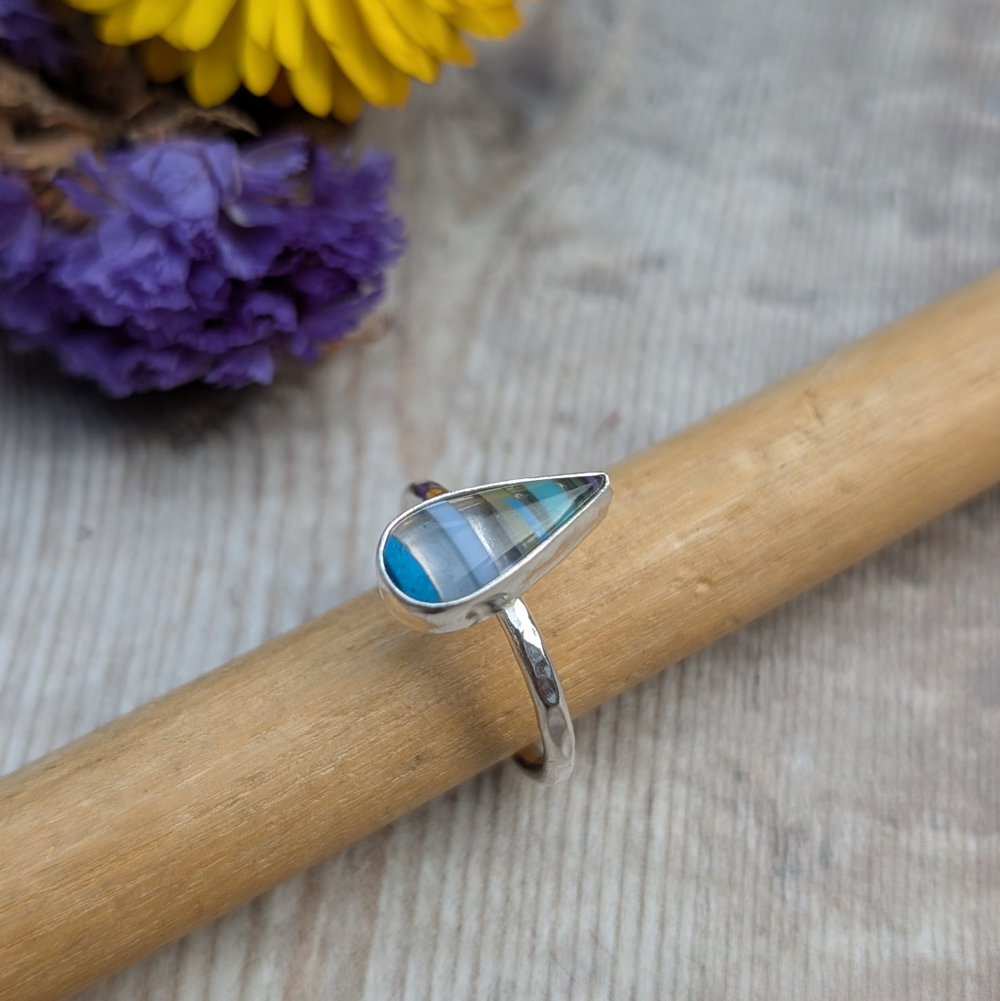 Close-up of silver teardrop Surfite ring resting on a wooden mandrel, with colourful flowers in the background.