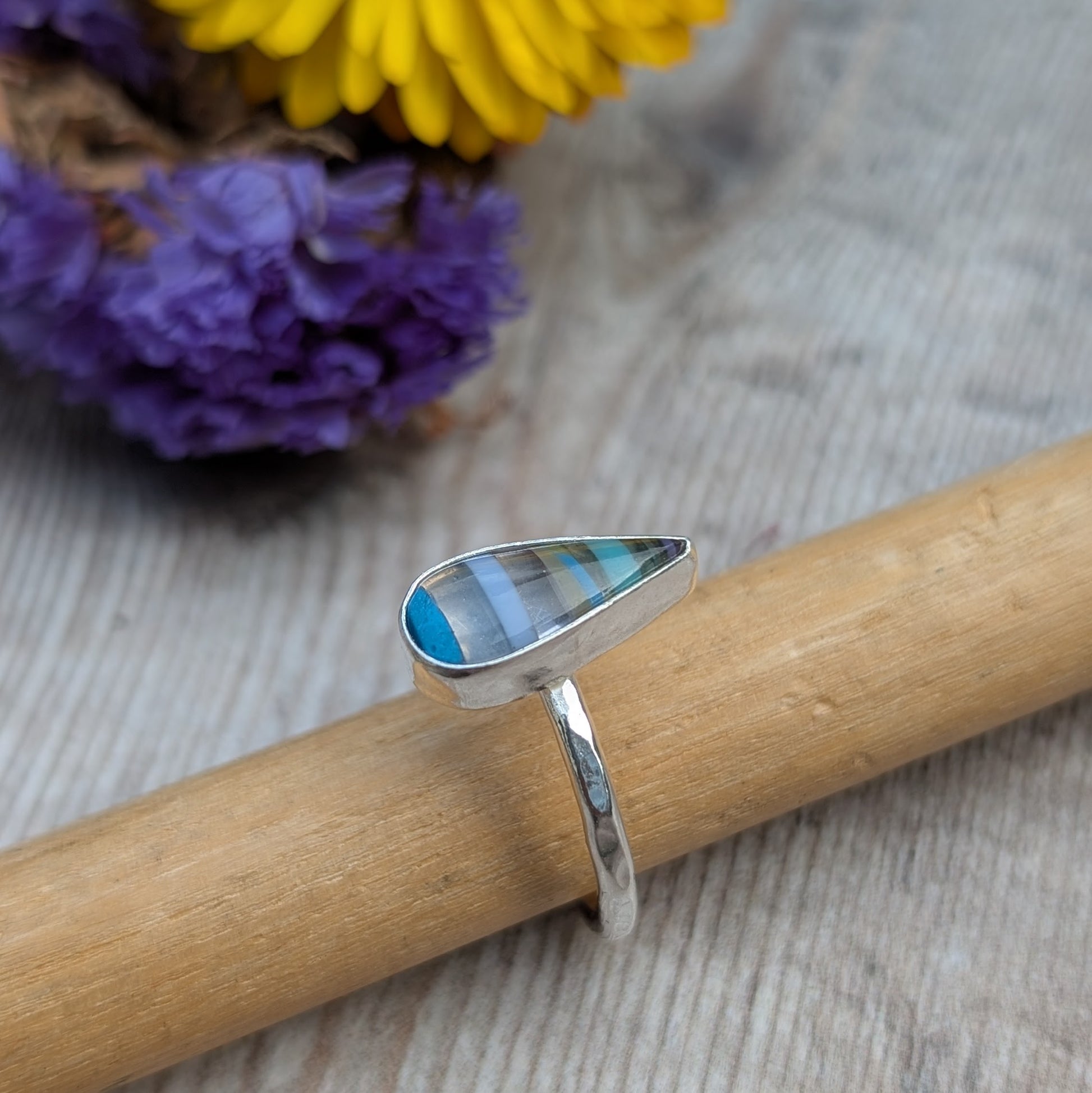Close-up of silver teardrop Surfite ring resting on a wooden mandrel, with colourful flowers in the background.
