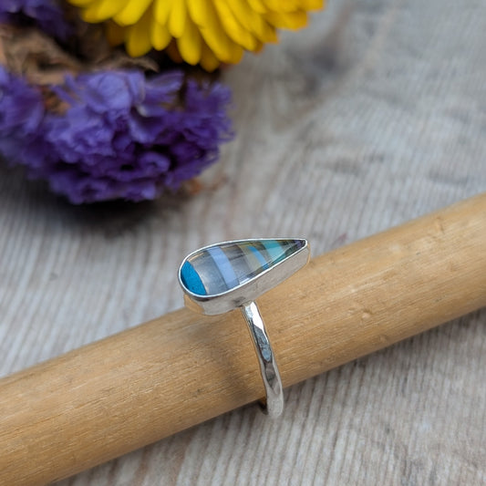 Close-up of silver teardrop Surfite ring resting on a wooden mandrel, with colourful flowers in the background.