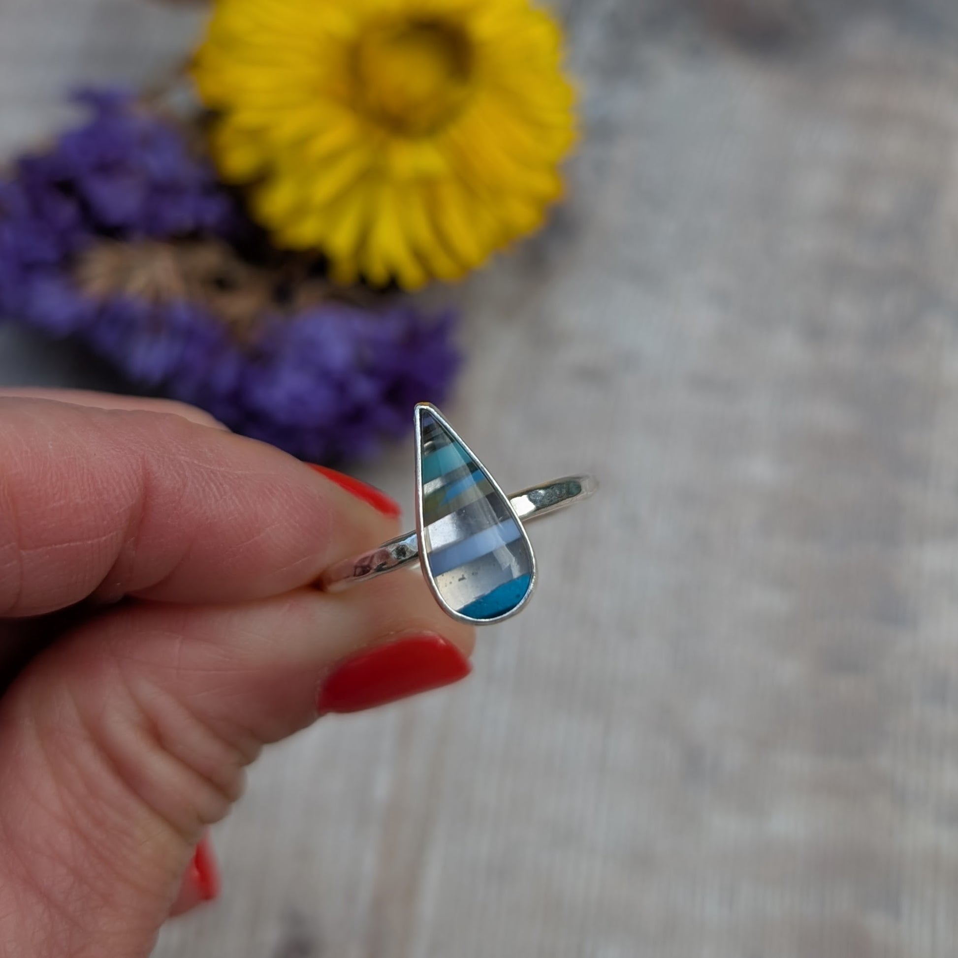 Handmade silver ring with teardrop-shaped Surfite stone in layered blue and white stripes, held between fingers with red nails.