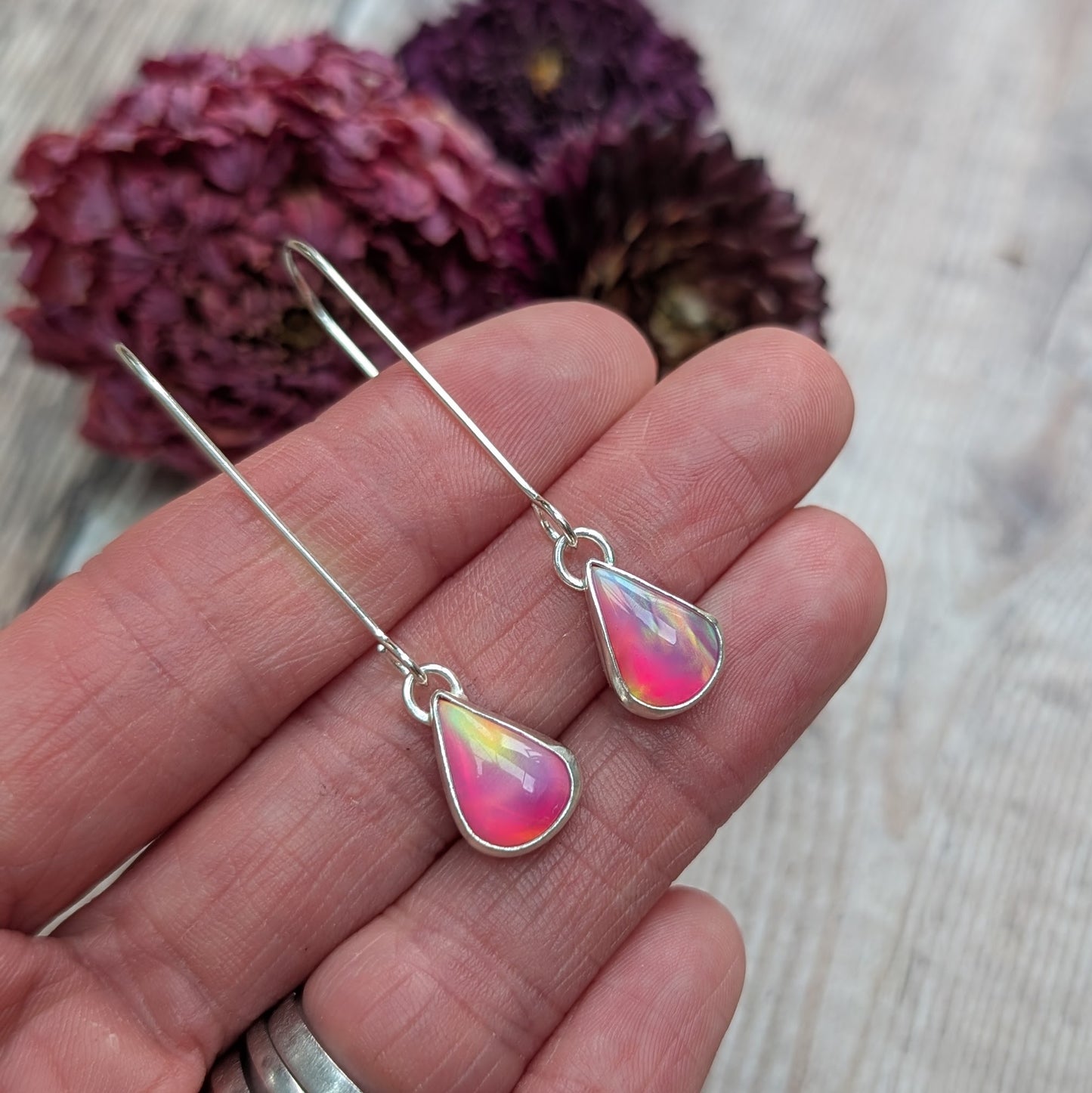 Close-up view of a hand holding a pair of Silver Teardrop Earrings with Pink Aurora Opal, showing their iridescent colours ranging from pink to yellow and hints of blue.