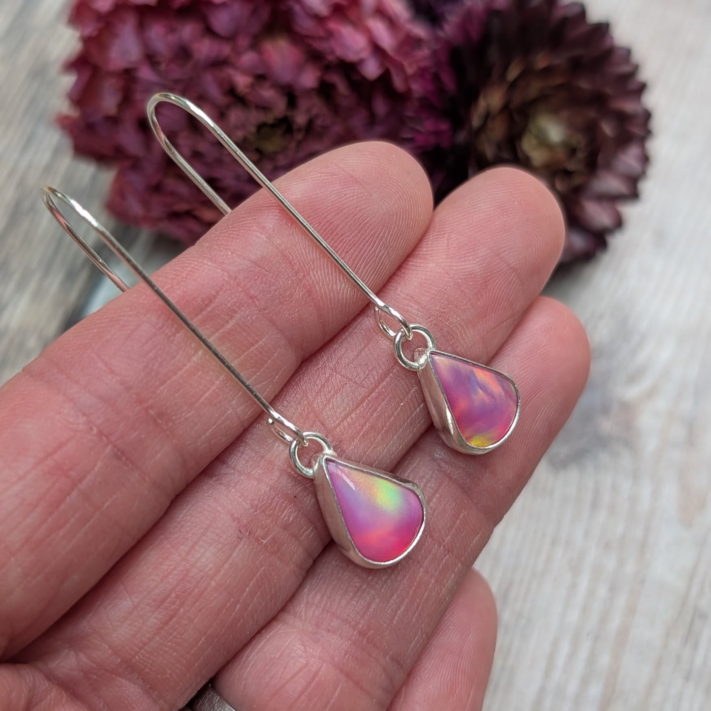 Close-up view of a hand holding a pair of Silver Teardrop Earrings with Pink Aurora Opal, showing their iridescent colours ranging from pink to yellow and hints of blue.