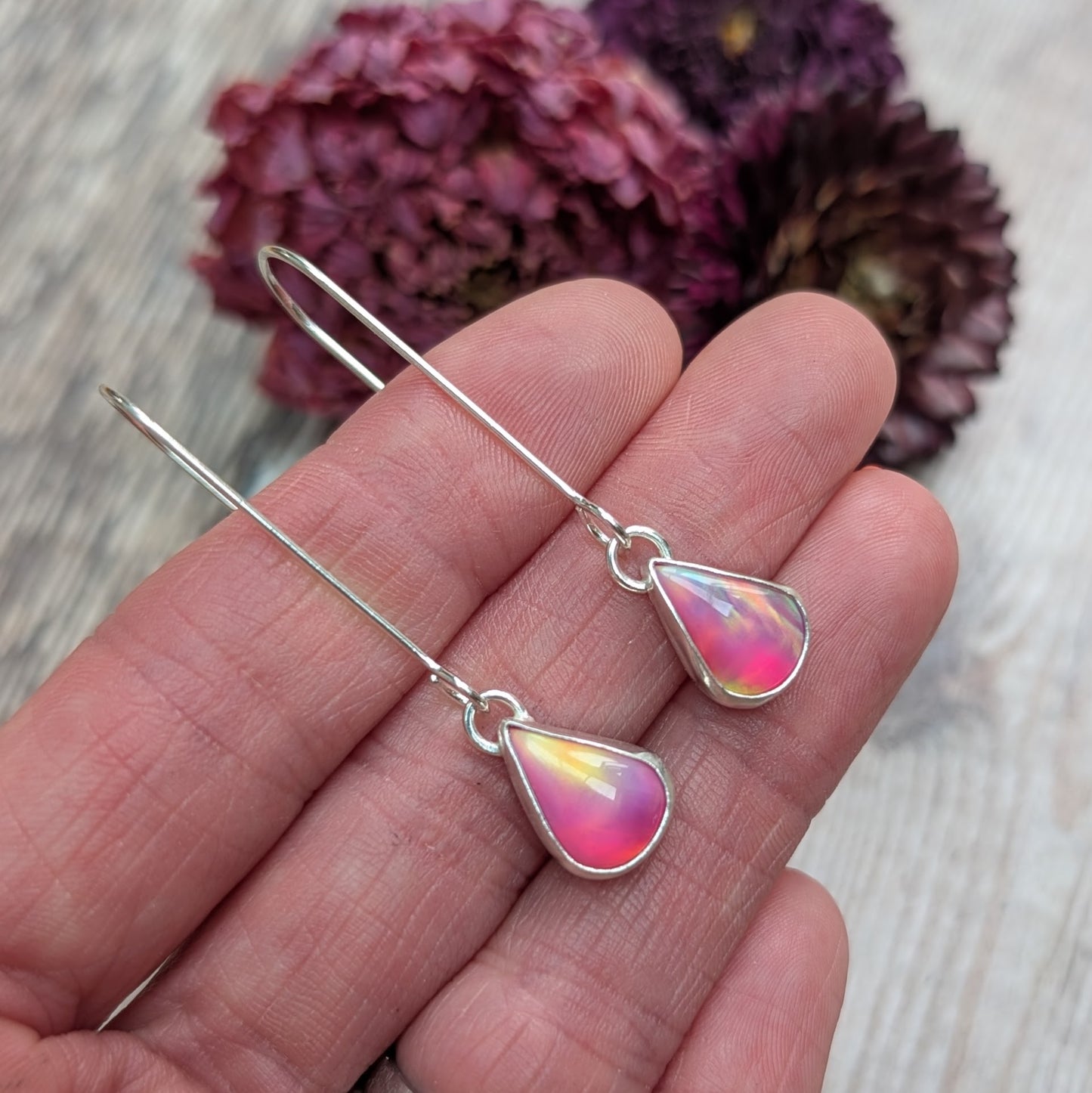 Close-up view of a hand holding a pair of Silver Teardrop Earrings with Pink Aurora Opal, showing their iridescent colours ranging from pink to yellow and hints of blue.