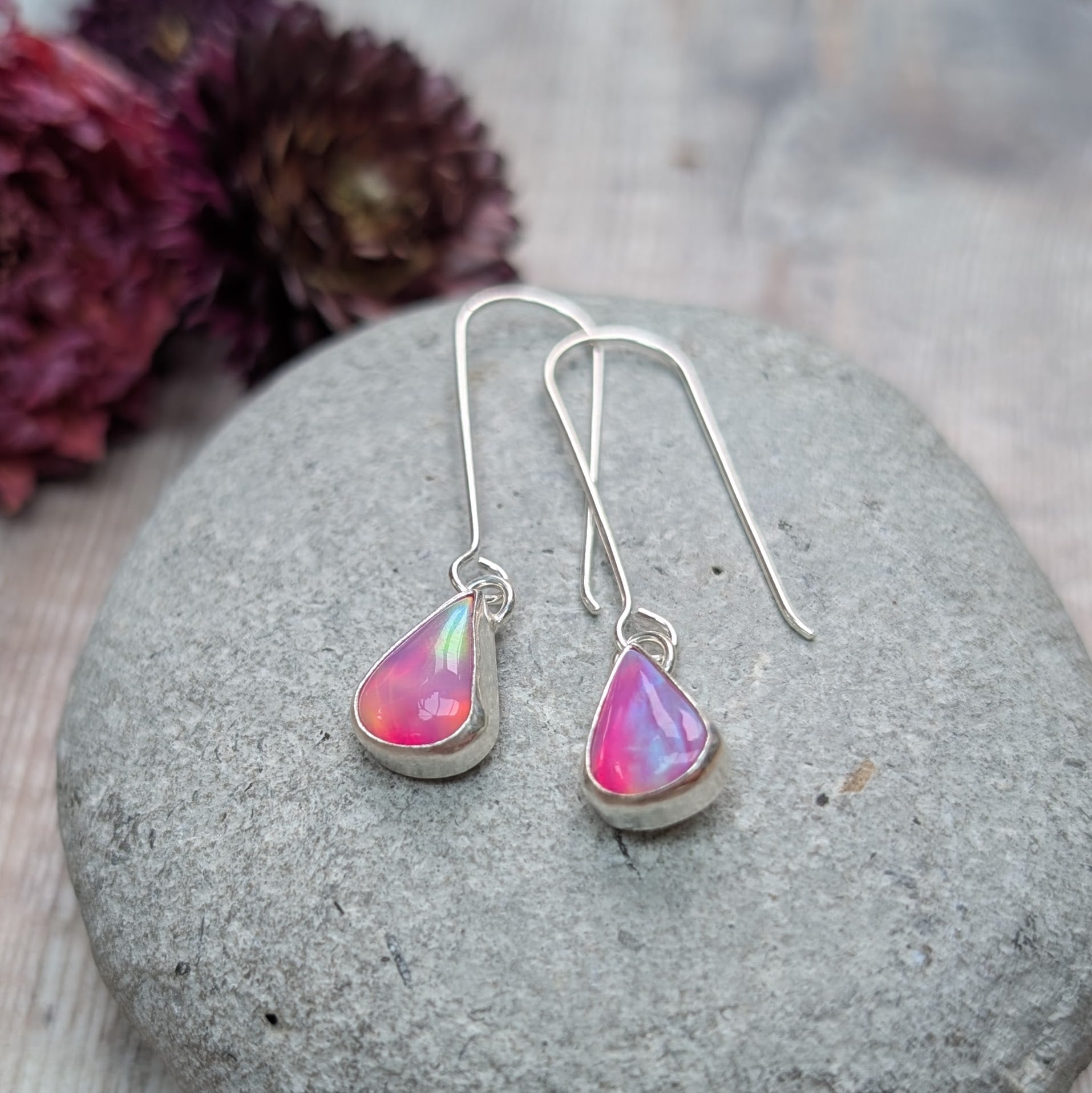 A pair of Silver Teardrop Earrings with Pink Aurora Opal displayed on a smooth grey stone, with soft-focus dried flowers in the background.