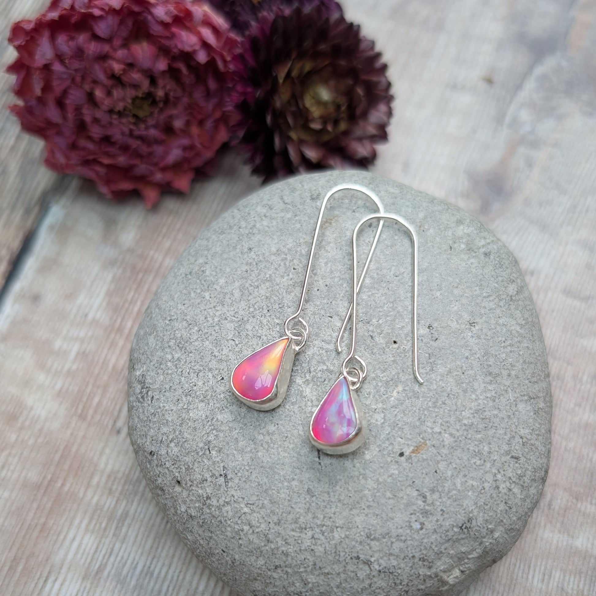 A pair of Silver Teardrop Earrings with Pink Aurora Opal displayed on a smooth grey stone, with soft-focus dried flowers in the background.