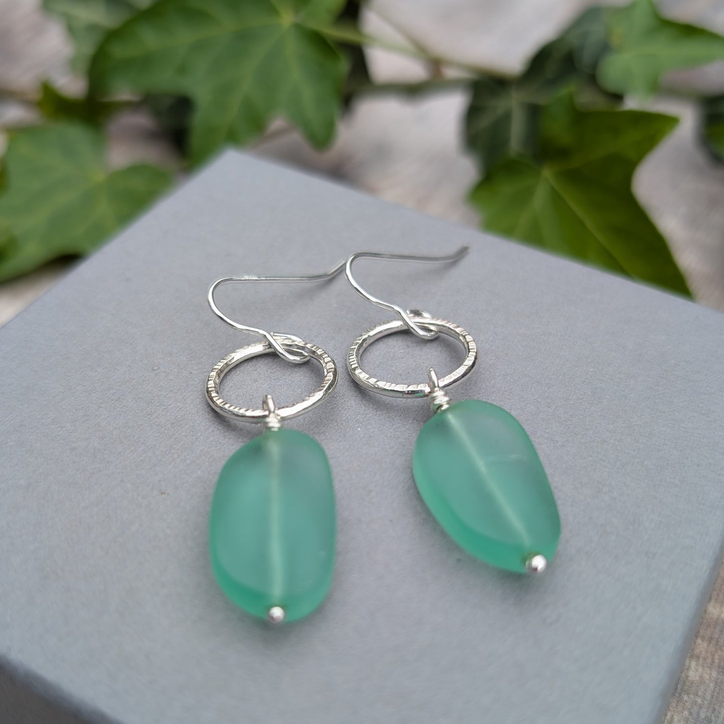 Silver Textured Circle Earrings with Aqua Sea Glass Beads displayed on a grey jewellery box with ivy leaves behind them.