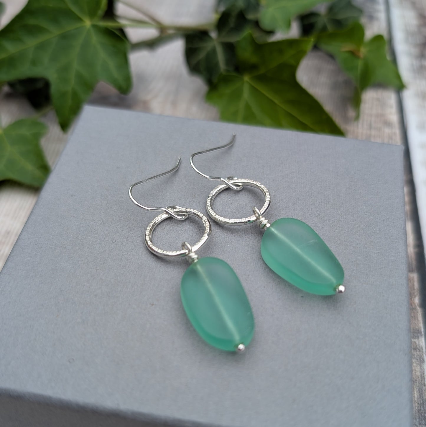 Silver Textured Circle Earrings with Aqua Sea Glass Beads resting on a grey jewellery box with green ivy in the background.