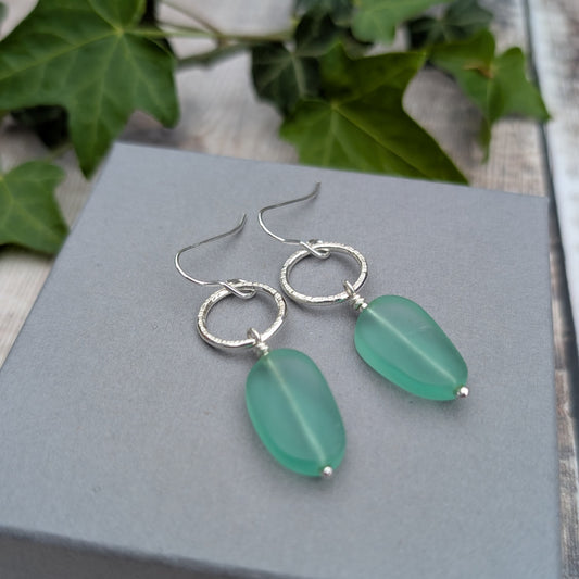 Silver Textured Circle Earrings with Aqua Sea Glass Beads resting on a grey jewellery box with green ivy in the background.