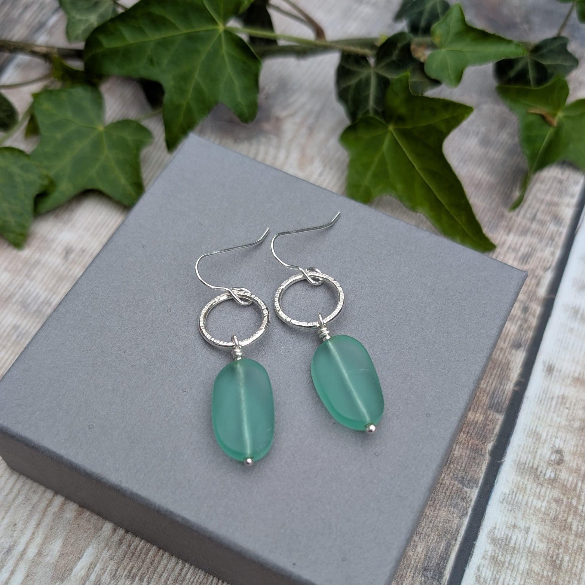 Pair of Silver Textured Circle Earrings with Aqua Sea Glass Beads displayed on a grey jewellery box outdoors.