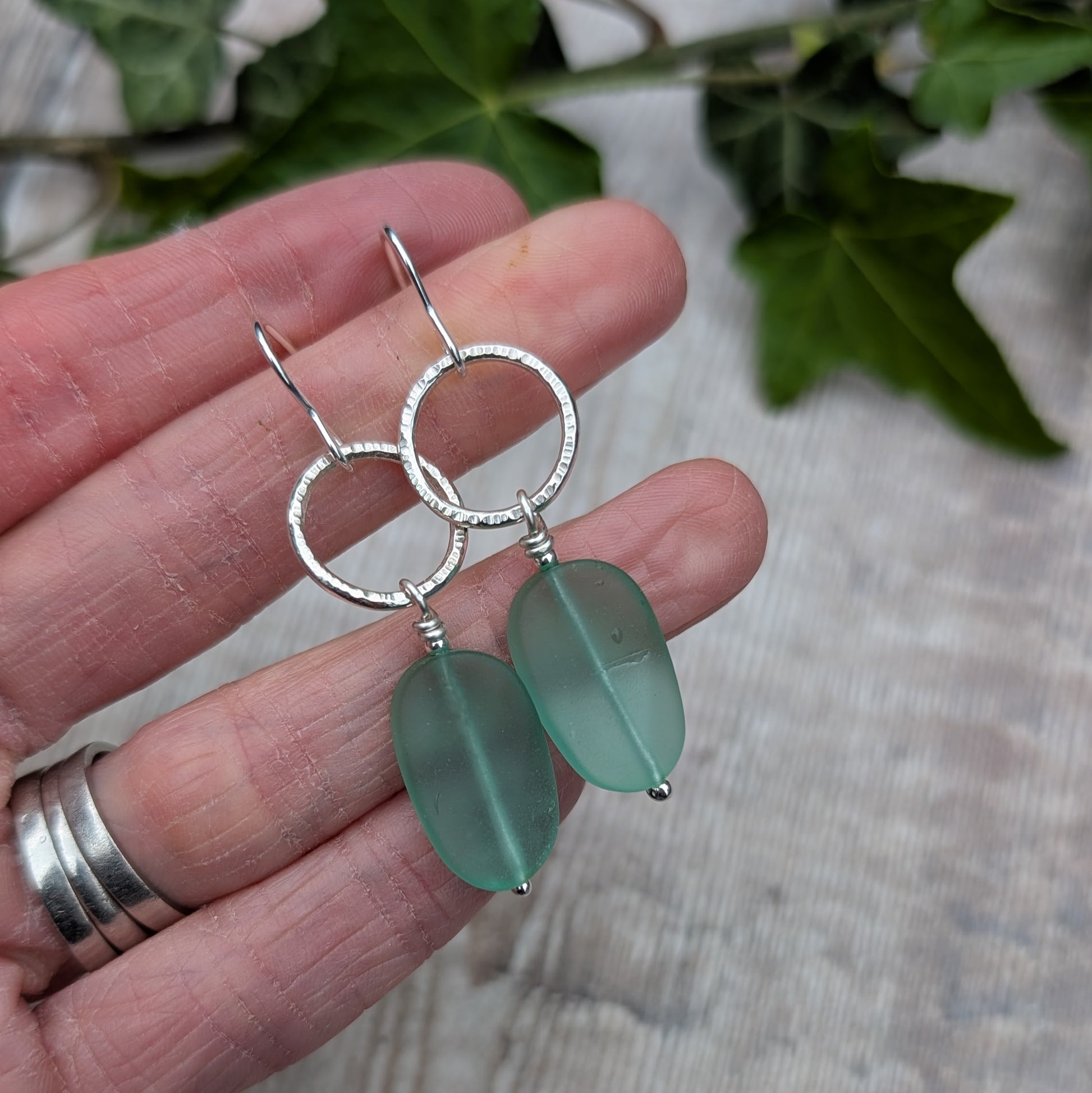 Handmade Silver Textured Circle Earrings with Aqua Sea Glass Beads shown against a wooden background while being held in a hand.