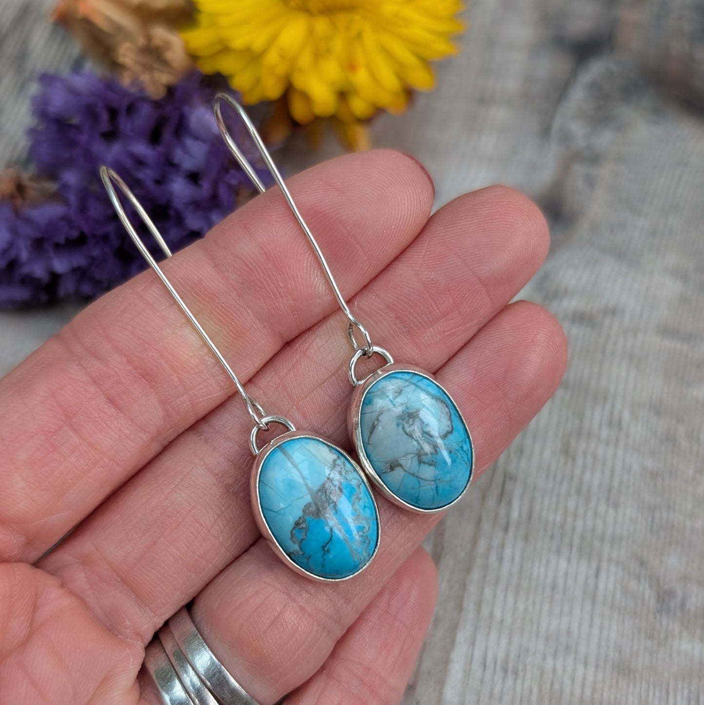 The handmade silver earrings held in a hand, highlighting the shape of the stones, their blue tones, and the smooth silver settings.