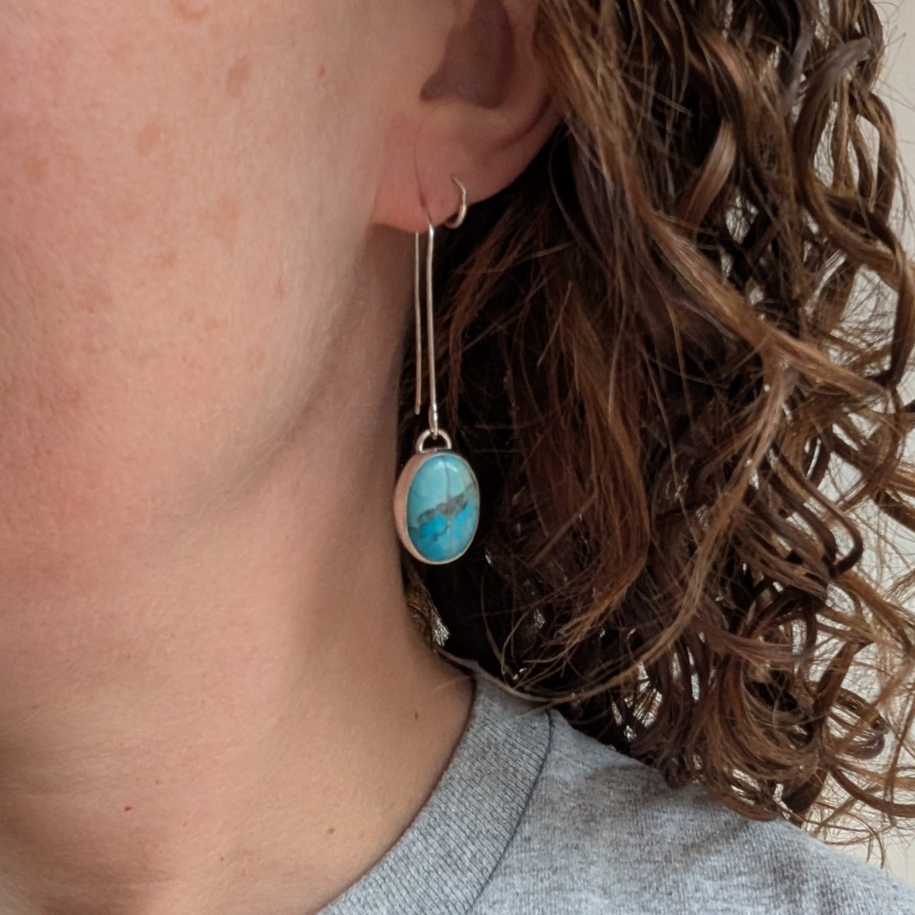 A close-up of a person wearing a long handmade silver earring featuring an oval Turquoise Howlite stone in a bezel setting. The earring hangs beside curly brown hair and a grey top is visible.