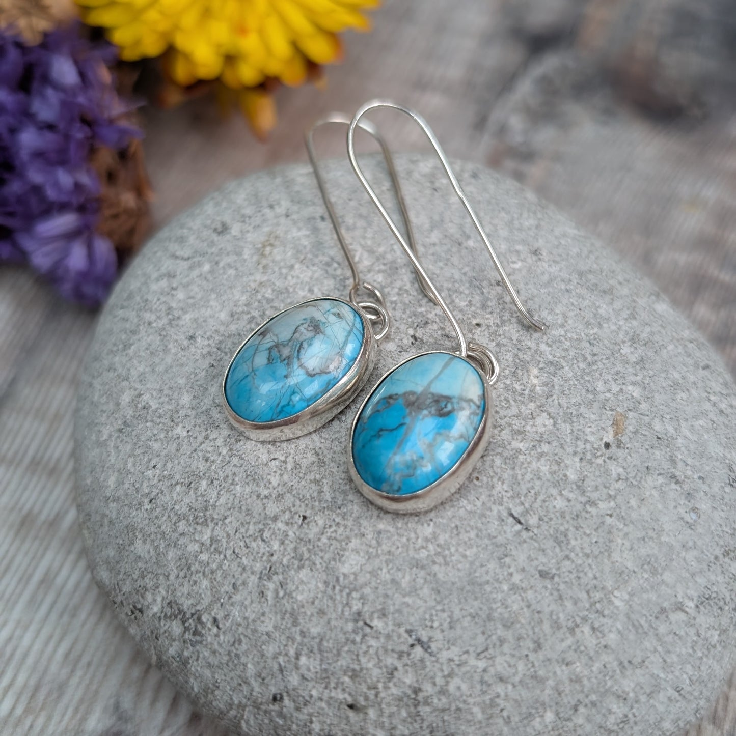 A pair of handmade silver earrings with oval Turquoise Howlite stones resting on a smooth grey pebble, with blurred yellow and purple flowers in the background.