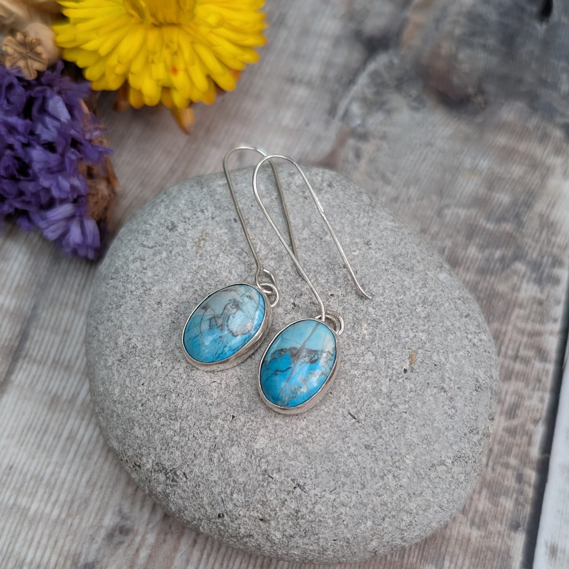 Long silver Turquoise Howlite earrings displayed on a grey pebble, showing the oval stones and simple handmade ear hooks.