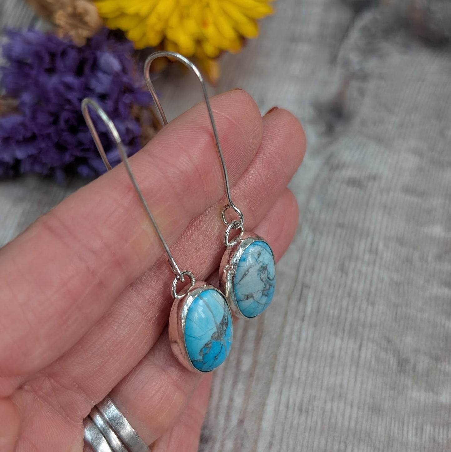 The earrings held gently in a hand, showing the long silver ear wires and oval Turquoise Howlite stones.