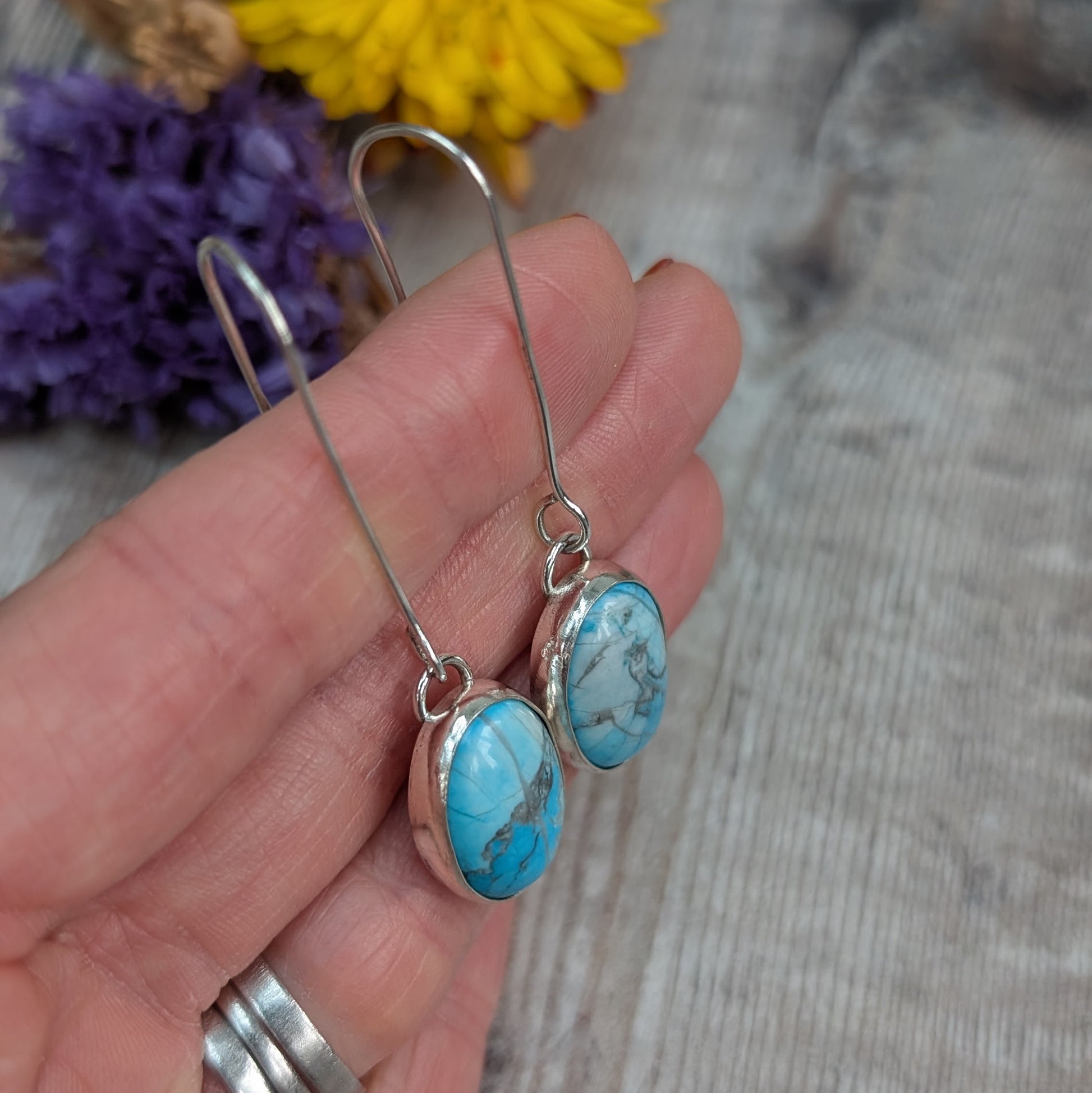 The earrings held gently in a hand, showing the long silver ear wires and oval Turquoise Howlite stones.