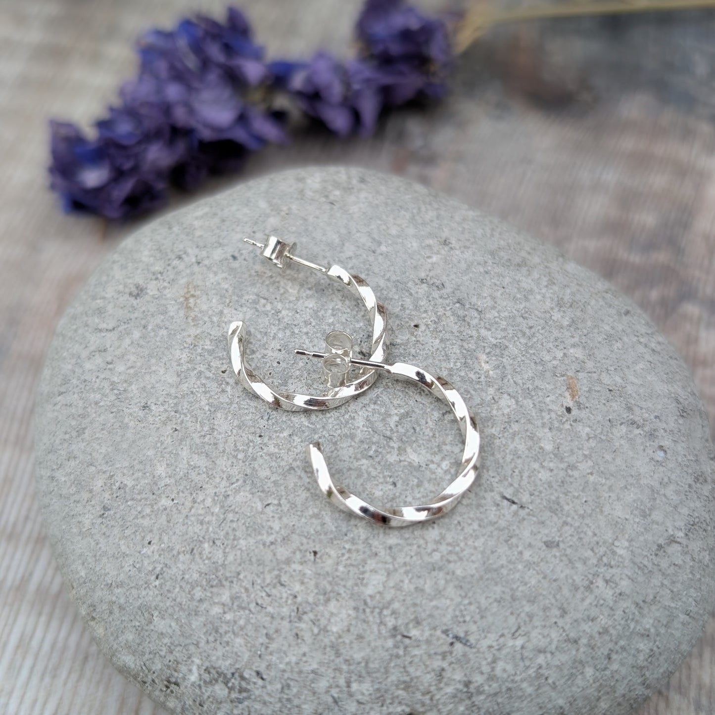 Handmade twisted sterling silver hoop earrings displayed on a grey stone, showing the curve and reflective surface.