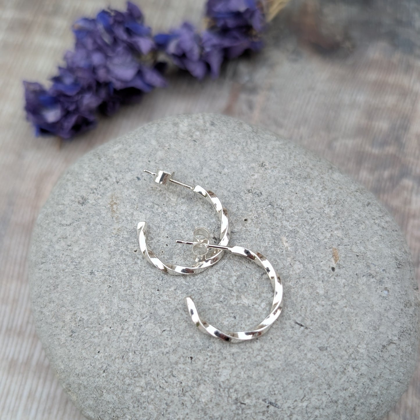 Pair of handmade twisted sterling silver hoop earrings resting on a smooth grey stone, with soft purple dried flowers in the background.