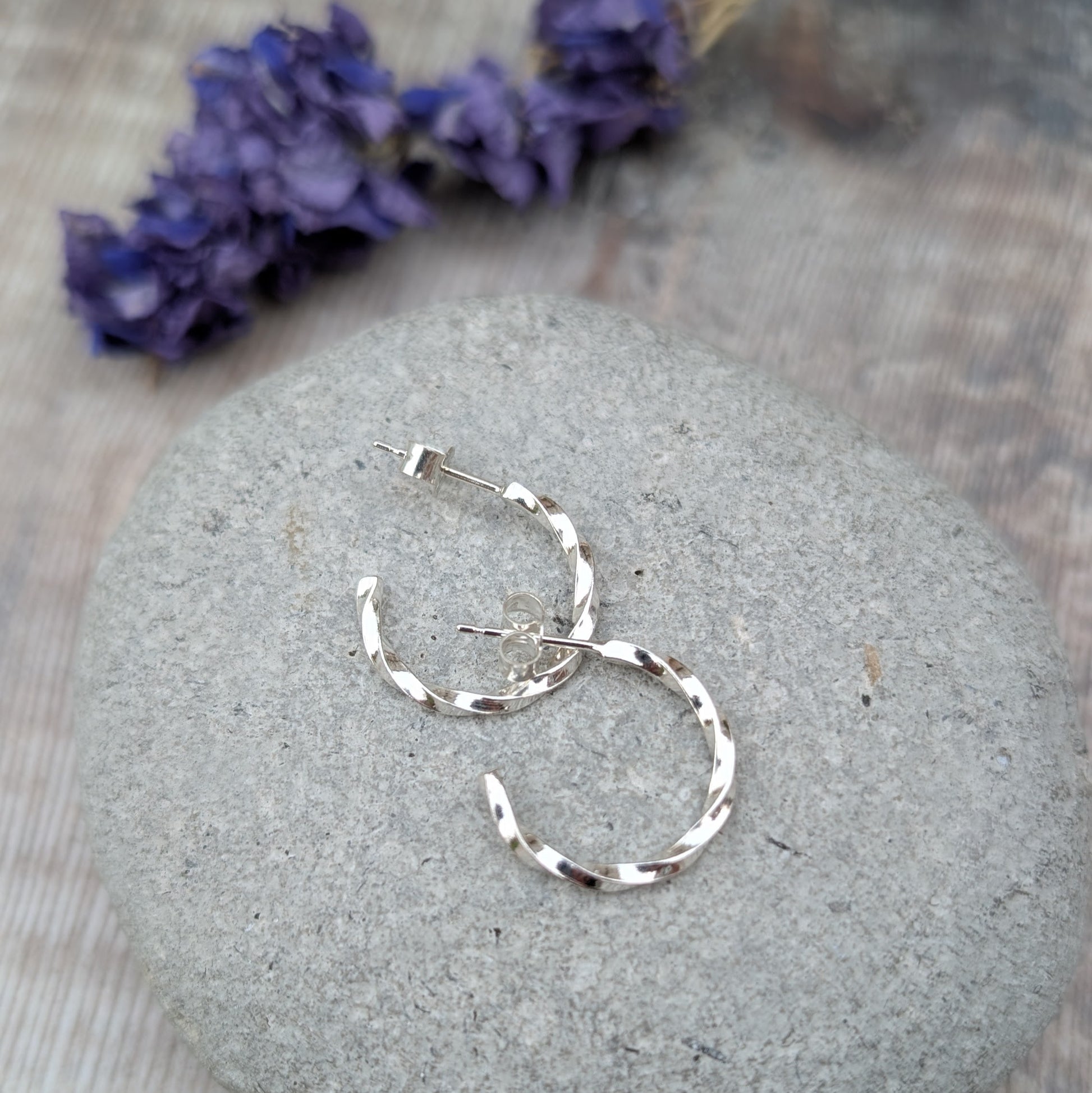 Pair of handmade twisted sterling silver hoop earrings resting on a smooth grey stone, with soft purple dried flowers in the background.